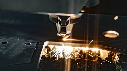 A machine lasers lines into a thick metal plate. Small orange/yellow sparks can be seen under the plate.