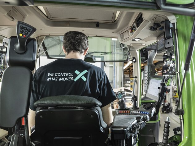 An employee of Arnold NextG can be seen from behind, through the rear window of a tractor cab. His T-shirt reads “We control what moves.” 